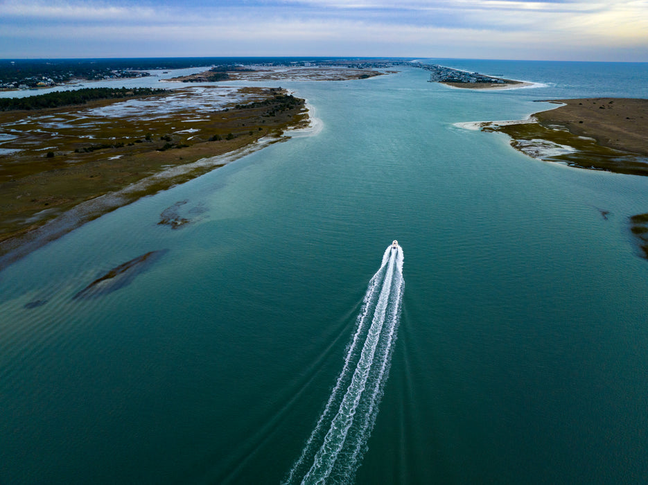 Aerial view of a narrow waterway between sandy and marshy islands, with a small white motorboat leaving a white wake through the turquoise-green water under a pale sky.