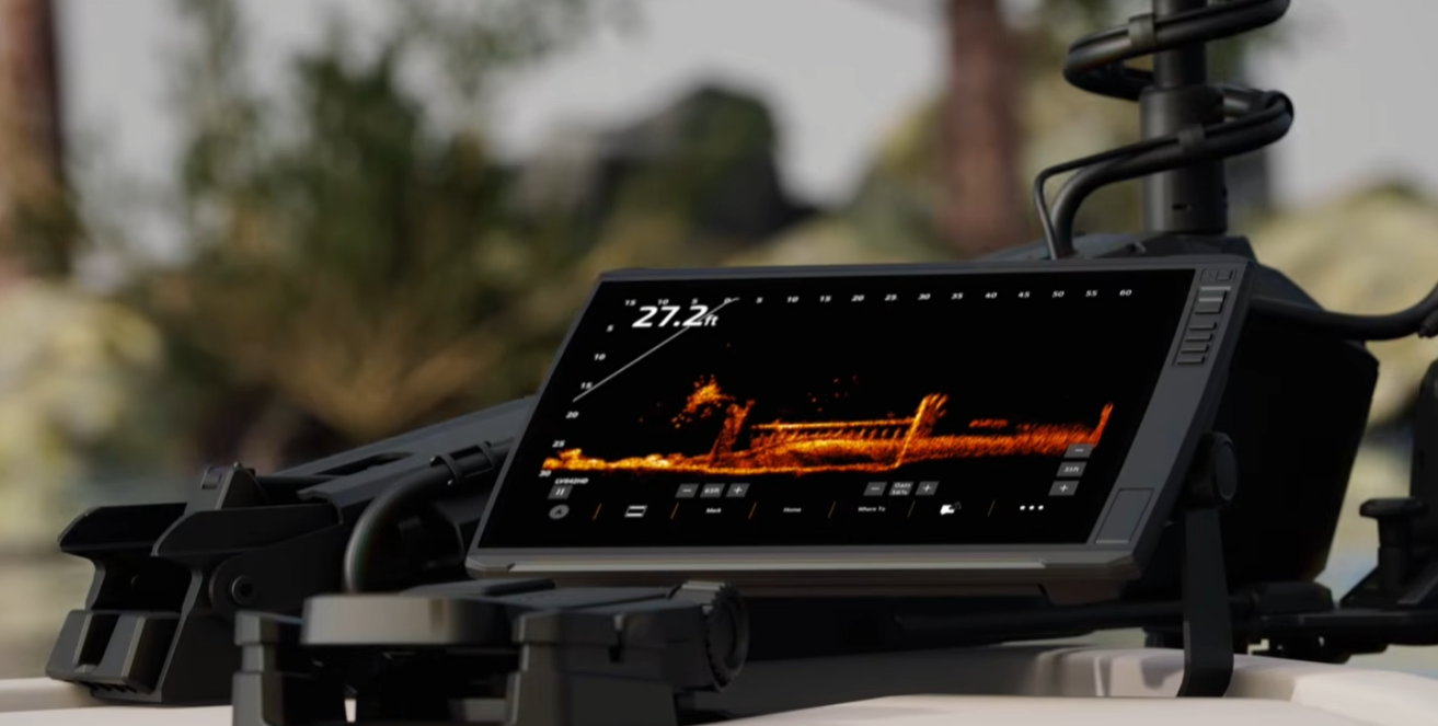 A black electronic fishfinder or sonar display screen mounted on a device, showing an orange and black underwater fish shape, with a blurred outdoor background.